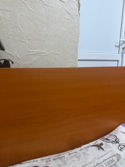 A close-up, eye-level, indoor shot shows a wooden surface in the foreground, possibly a desk or a bed frame, with a slightly textured, light-colored wall in the background. To the right of the wall is a white door or window frame with a white handle and lock. A small red speck is visible on the wooden surface. The bottom right corner of the frame reveals a portion of a patterned fabric, possibly a pillow or blanket, with text and decorative elements including a birdcage and a silhouette of a bird. The scene is devoid of human presence. The lighting is even and diffused.