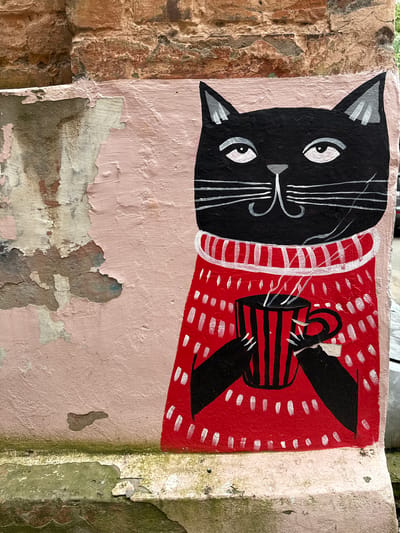 The image is a close-up photograph of a mural painted on a weathered, pink-colored concrete wall. The mural depicts a black cat wearing a red, high-necked sweater with white dashes. The cat has white eyes, white whiskers, and a grey nose. It is holding a red and black striped mug with steam rising from it. The concrete wall is cracked and peeling in places, revealing older layers of paint and a rough, textured surface underneath. Above the pink wall is a section of rough, reddish-brown brickwork. The bottom of the image shows more peeling paint and some green mossy growth on the lower concrete structure. The style of the mural is simplistic and cartoonish. The specific location in the city of Votkinsk, Russia is not determinable from the image itself.