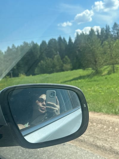 The image shows a car's side mirror, reflecting a person taking a photo with a smartphone. The person is wearing sunglasses. The mirror is attached to a car moving along a road. The background, visible through the car's window and partially reflected in the mirror, consists of trees, a green field, and a blue sky with clouds. The location is identified as being somewhere in the city of Staryi Chul'tem, Russia, based on the image's context (although this cannot be determined from the image itself).