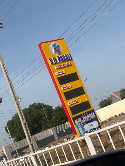 A tilted, medium shot captures a yellow gas station sign with red trim against a clear blue sky. The sign displays the company name "S.D. PASALI Oil & Gas Nig. Ltd." prominently, with smaller labels for "PETROL," "DIESEL," and "LPG" above black price displays. A graphic featuring an oil barrel and the text "LPG" is visible at the bottom of the sign. In the background, green trees and a light-colored building with a darker band are partially visible. A concrete utility pole with wiring stands to the left of the sign, and a white metal fence runs across the foreground. Text on a nearby wall reads "NO SMOKING." The scene appears to be an outdoor location in the city of Rahwol Chom, Nigeria.