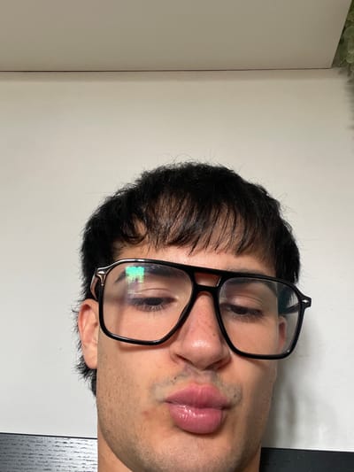 A close-up portrait features a person, likely male, positioned vertically on their right side, against a plain, light-colored background. The frame captures the individual's head and the upper part of their shoulders. They have dark, short hair, which frames their face, with some strands falling on the forehead. The person is wearing large, black-framed glasses.
Their eyes are partially closed or looking downwards, visible through the lenses of the glasses. A distinct reflection of green, square-shaped pixels is present on the right lens, possibly from a screen or artificial light source. The person's lips are prominent and appear slightly parted or in a relaxed pout. There's visible stubble on the chin and upper lip area. The right ear is partially visible, protruding from beneath the hair.
The background is predominantly a smooth, off-white wall or surface, with a hint of a darker, textured surface (possibly a table) visible along the right edge of the frame. In the very top left corner, a soft green blur suggests foliage, potentially from an indoor plant. The lighting appears soft and even, highlighting the facial features without harsh shadows. No text is visible in the image. The overall impression is a candid or posed close-up portrait, focusing on the individual's face and features.