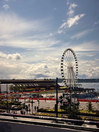 A Ferris wheel dominates the skyline, positioned near a waterfront. The ground view reveals a pedestrian area with people walking, alongside commercial buildings and streetlights. The background features a body of water, possibly a bay or lake, with land visible in the distance. The sky is filled with wispy clouds, suggesting a partially cloudy day.