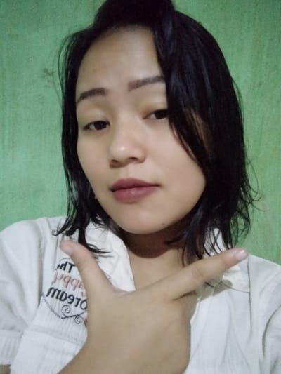 A young Asian woman poses indoors with a green wall background. She has short, dark hair and wears a white collared shirt with text visible on the chest. Her makeup includes defined eyebrows and lip color. She's holding up her hand, index finger extended, pointing to the right. Her expression is neutral, looking towards the camera. The lighting in the image is diffused, with slight shadows across her face.