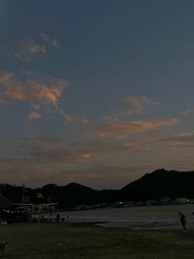 This image captures a beach scene in San Juan del Sur, Nicaragua, during twilight. The sky displays a gradient of deep blue at the top, transitioning to lighter hues with scattered clouds tinged with pink and orange, indicative of sunset. In the foreground, a sandy beach stretches out, with a few dark figures walking or standing. To the left, a thatched-roof structure with string lights suggests a beachfront establishment. Beyond the beach, the ocean is visible with gentle waves lapping the shore, and further back, a line of buildings and homes are nestled against the dark, tree-covered hills that dominate the background. A tall communication tower is also visible on one of the hills. The overall impression is one of a serene evening by the coast.