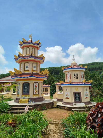 The image shows two ornate, multi-tiered stupas in a cemetery in Xuân Sơn, Vietnam. The stupas are painted in pale yellow and decorated with intricate gold dragons, blue trim, and red accents. Chinese characters and Buddhist swastikas adorn their surfaces. The larger stupa on the left has multiple levels and is topped with a golden swastika symbol. The smaller stupa on the right is also decorated with Chinese characters and a swastika. Both stupas have plaques or inscriptions on their facades, likely commemorating individuals. The surrounding area is lush with green vegetation, including bushes, trees, and some flowering plants. In the background, a verdant hillside covered in trees slopes upwards. The sky is bright blue with scattered white clouds. The overall scene suggests a serene and well-maintained burial ground.