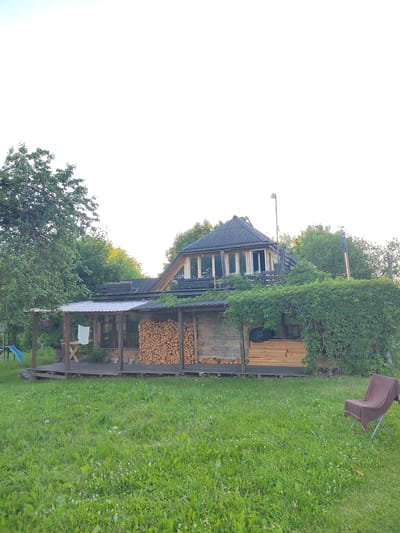 The image depicts a rustic wooden house situated in a grassy, rural setting, likely in the Lyntupsky Selsovet, Belarus. The house features a covered porch with a corrugated metal roof, supported by wooden posts. Piles of firewood are neatly stacked under the porch, suggesting a dwelling that relies on wood for heating or cooking. Green ivy or other climbing plants are abundant, growing over parts of the house and its upper levels, creating a natural, overgrown aesthetic.
The house itself has a multi-level design with visible wooden plank construction and a dark-colored, sloped roof. The upper section has large windows, offering a view and possibly serving as an attic or an additional room. A light fixture is mounted on a pole above the house.
In the foreground, a lush green lawn is dotted with small white flowers, indicating a natural or unmanicured yard. To the right, a foldable camping chair draped with a brown blanket sits on the grass, suggesting a place for relaxation or observation of the surroundings. To the left, a blue slide and a portion of a playground structure are visible, hinting at the presence of children or a recreational area.
The sky is bright and overcast, suggesting a daytime scene, possibly late afternoon or early evening, given the soft light. The overall impression is one of a peaceful, somewhat wild, and self-sufficient rural dwelling. There is no visible text in the image.