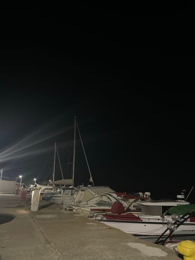 In Limenas Avdiron, Greece, at night, several boats are docked in a harbor. The scene is illuminated by artificial lights. In the foreground, a concrete walkway runs along the edge of the harbor. A yellow bollard is visible on the right side of the walkway. Several boats are moored alongside the walkway, including a white motorboat with red seating and a green cover. Further back, two sailboats with tall masts are visible. The sky is dark, with some light beams from unseen sources cutting through the darkness. The overall impression is of a quiet harbor at night.