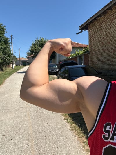 A person's flexed arm and bicep are prominently featured in the foreground. The individual is wearing a red basketball jersey with white and black trim, and the letters "SA" are visible on the jersey. The background depicts a sun-drenched rural street in the village of Konop, Bulgaria. A dirt road curves to the left, bordered by green grass and trees. Several houses with red-tiled roofs and brick facades are visible, as are two black cars parked on the right side of the road. A power line with poles stretches across the blue sky. The overall scene suggests a warm, clear day in a village setting.
