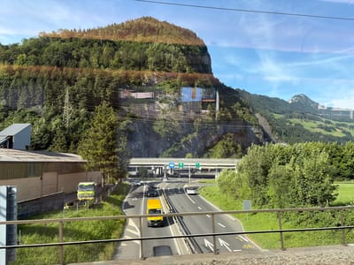 This image captures a view from a moving vehicle, likely a train, traveling along a highway in a mountainous region. The highway, a multi-lane asphalt road, is populated by several vehicles including cars, a yellow van, and a yellow truck. The truck appears to be unloading a dark material into a yellow bin. The road is bordered by grassy verges and lush green trees. A pedestrian bridge crosses over the highway in the middle distance. In the background, a steep, tree-covered mountain rises dramatically, with some rock faces visible. The sky above is blue with scattered white clouds. Reflections from the vehicle's windows are visible throughout the image, distorting the scene slightly.

While the image provides a scenic overview of a mountainous landscape and a highway, it does not contain specific visual cues that definitively place the location within the city of Schwyz, Switzerland. The general topography suggests a location in the Swiss Alps, which would be consistent with Schwyz, but further information would be needed for precise identification.