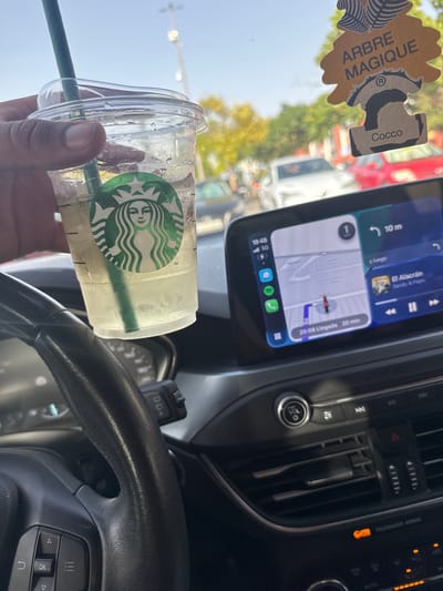 In the foreground, a hand is holding a clear Starbucks iced beverage with a green straw, obscuring the bottom of the image. The car's interior is visible, including the steering wheel with control buttons on the left. The infotainment screen displays a navigation map with a route highlighted, indicating an estimated arrival time of "20:08 Llegada 20 min." Music playback is also shown on the screen, with the song "El Alacrán" by Sandy & Papo. A tree-shaped air freshener labeled "ARBRE MAGIQUE COCCO" hangs from the rearview mirror. The background shows a street scene with trees and cars, suggesting the vehicle is in motion or stopped in traffic. The overall setting appears to be a sunny day in a residential or commercial area of Arroyomolinos, Spain.