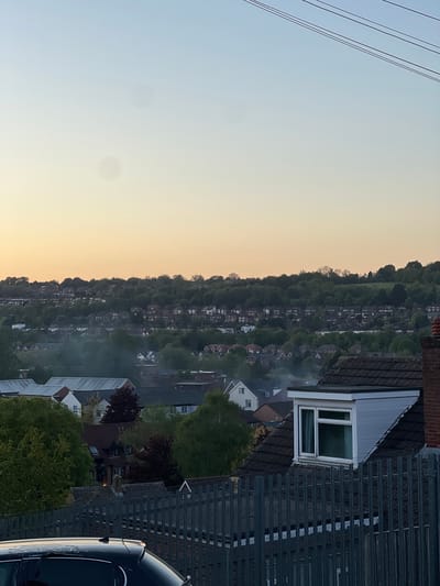 This image presents a panoramic view of a residential area at what appears to be either sunrise or sunset. The sky, which covers the upper portion of the frame (when oriented correctly), transitions smoothly from a pale blue overhead to a warm yellow and orange near the horizon, indicating clear weather. Several dark power lines traverse the top-left corner of the sky. Two faint, circular smudges, possibly dust on the camera lens, are visible in the expanse of the sky.
In the midground and background, a long, undulating hill covered with dense green trees stretches across the width of the image. Numerous residential houses with varying roof types and light-colored facades are nestled among the trees, extending far into the distance along the hillside, suggesting a suburban setting like High Wycombe.
The immediate foreground on the right side features the partial view of a dark blue or black car, showing its hood, windshield, and roof. Some faint, illegible white text or markings are visible on its side panel. Adjacent to the car and further left, several houses are visible, featuring both dark grey corrugated roofs and traditional reddish-brown tiled roofs. One house prominently displays a white dormer window. A small yellow triangular sign with a black warning symbol (indistinguishable) is attached to one of the grey corrugated roofs. Various trees with green and reddish-brown foliage are scattered amongst the houses. There is no visible human activity or specific event taking place.