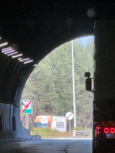 This media file captures a view from inside a dark, enclosed space, possibly a vehicle or tunnel, looking outwards. The scene outside is dominated by lush green foliage, suggesting a natural setting such as a roadside or park. A white line, likely a road marking, stretches across the frame, indicating a pathway or road.
Several objects are visible in the background. A traffic sign with a red diagonal line on a white circle within a green square is present, signifying a prohibition. Further into the scene, a white rectangular object with dark lettering, possibly a sign or a container, is partially obscured by the foliage. A light-colored vehicle, with orange accents, is also visible.
At the top of the frame, a digital display shows red circular lights, likely part of a vehicle's indicator or warning lights, and some unreadable digital text. A small black object, possibly a rearview mirror, is mounted near the top.
The lighting suggests it is daytime, and the presence of dense foliage implies it might be spring or summer. The overall mood is somewhat obscured by the dark foreground and the slightly blurry nature of the image, but the outward view conveys a sense of moving through or observing a relatively serene, green environment. The text visible on the sign is "MAERSK".