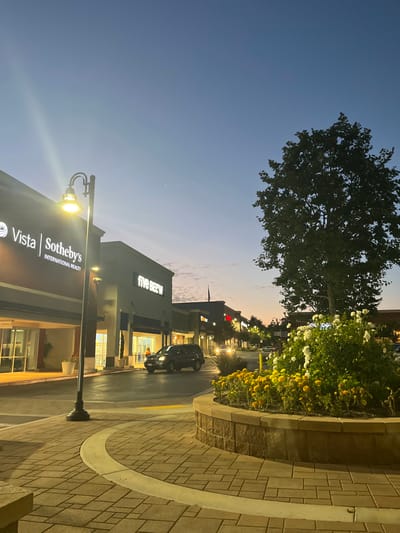 This image depicts a commercial plaza at dusk or early evening in the Peninsula Center, Rolling Hills Estates, United States. The sky exhibits a gradient from dark blue overhead to lighter blue and faint orange near the horizon, with two bright celestial objects visible in the upper-left quadrant.

In the foreground, a brick-patterned pedestrian walkway or plaza section is visible. A tall, black streetlamp on the left emits a bright yellow light, casting a glow. To its right, a raised stone-faced planter contains dark green foliage with white blossoms and smaller plants bearing yellow-orange flowers. Behind the planter, a large tree with dense green leaves is silhouetted against the sky.

In the midground, an asphalt drive or roadway is present. A dark-colored SUV is in motion, oriented towards the right, with its rear section visible. Further down the road, the headlights of another vehicle are discernible. Commercial buildings line the street, with illuminated signs. Prominently featured on the left building is "Vista | Sotheby's International Realty," and further right, "FIVE BELOW." These buildings are light-colored with various architectural features and multiple light sources.

No individuals are visible within the frame. Artificial lighting is active from the streetlamp, building signs, and interior spaces, indicating nighttime operations.