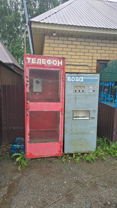 This image captures an outdoor scene in the village of Bol'shoi Mashlyak, Russia, featuring two distinct vending machines situated near a building. On the left is a weathered red telephone booth, its glass panels revealing an old rotary dial phone mounted inside. The booth shows signs of age, with peeling paint and visible wear. To its right stands a pale blue vending machine, labeled in Russian with "ВОДА" (Water) and "газированная" (sparkling). This machine has a dispenser slot with a glass cup inside, and a control panel with buttons and small displays. Both machines are positioned on a gravel surface, with some weeds and greenery growing around their bases. The background includes a building with yellow brickwork and a corrugated metal roof, as well as a dark brown metal fence. The overall impression is of an older, possibly disused, public utility area.