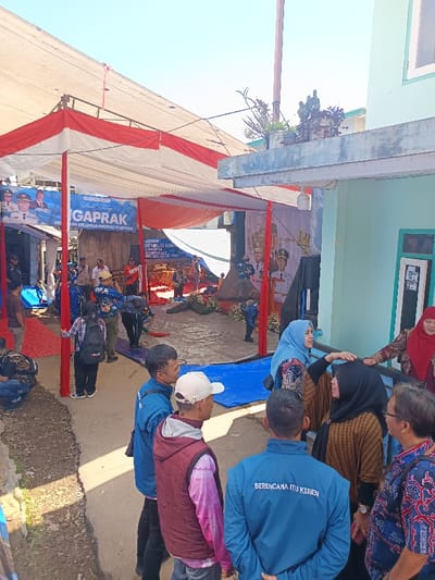 An outdoor community event is underway in Kawungluwuk, Indonesia, under a large temporary canopy made of white fabric with red and white accent strips. The canopy is supported by visible red poles. On the right, a light blue building features a flat roof with potted plants, a window, and a door. The ground in the immediate foreground is unpaved dirt, while red carpets and blue tarpaulins are laid out under the canopy.
Multiple individuals are present. In the foreground on the right, a group of approximately seven adults is engaged in conversation. This group includes a man in a blue long-sleeve shirt with "BERENCANA ITU KEREN" on the back, facing away; a man in a maroon vest and white baseball cap; another man in a blue jacket; a man in a patterned batik shirt and glasses; and three women. Two women wear hijabs – one in light blue, touching the head of a woman in a black hijab and brown patterned shirt; the third woman, wearing a red hijab and patterned shirt, is partially visible behind a railing.
Under the canopy, numerous other people are standing or moving. Visible subjects include individuals in blue shirts, some carrying backpacks, and others in various casual and patterned attire. Event banners are displayed, one on the left reading "Selamat Datang GAPRACK" and referencing "KELUARGA BERISIKO STUNTING" (families at risk of stunting), featuring images of officials. Another banner on the right mentions "REMBUG WARGA" (community discussion) and also displays images of officials. Decorative elements, possibly floral arrangements, are visible near the center of the event space under the canopy. The sky is clear and blue, indicating a bright, sunny day.