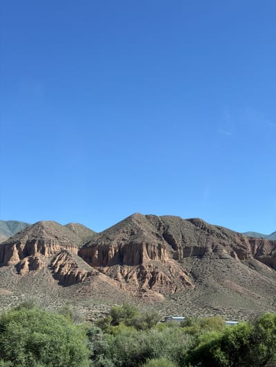 This media file captures a dramatic landscape of arid, mountainous terrain meeting lush vegetation under a clear blue sky. The image is oriented vertically, with the sky occupying the left half and the mountainous slope and trees on the right.
The primary subjects are the imposing, eroded rock formations that dominate the right side of the frame. These mountains display distinct layers and textures, indicative of significant geological history, with deep gullies and weathered surfaces. They are sparsely covered with low-lying green scrub, suggesting a harsh, dry climate.
On the far right, a dense growth of vibrant green trees and shrubs forms a stark contrast to the arid mountains. These trees appear healthy and full, hinting at a localized source of water, possibly a riverbed or spring, that supports this pocket of greenery. A thin, light-colored road or path is partially visible winding along the base of the trees, suggesting human passage through this rugged area.
The sky is a flawless, deep blue, with no clouds visible, indicating a bright, sunny day. The lighting suggests it is likely midday or early afternoon, given the strong illumination and the absence of long shadows.
The overall scene evokes a sense of natural grandeur and the stark beauty of a remote, mountainous region. The juxtaposition of the dry, ancient rock with the vibrant, living foliage creates a visually striking composition. The location context provided, Kok Moynok Eki, Kyrgyzstan, aligns with the visual cues of a high-altitude, arid environment with potential for localized oases. There is no visible text in the image.