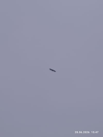 The image shows a clear sky with a gray, hazy atmosphere. A small, dark, elongated object is visible in the center of the frame, appearing to be airborne. The object is indistinct and could be a drone or a bird. The timestamp "28.06.2026 15:47" is present in the bottom right corner, indicating the date and time the photograph was taken. The location is Okhotskoe, Russia, though no specific urban or environmental features are discernible in the image.