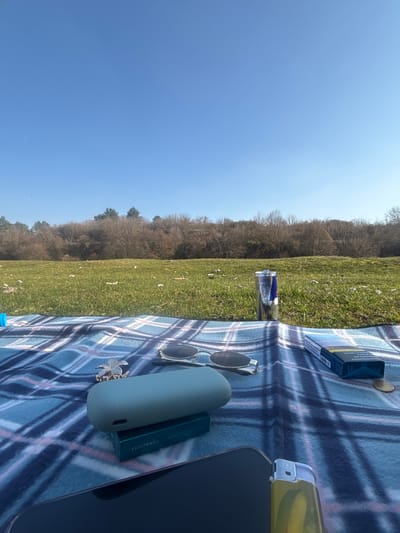 The scene is an outdoor setting located in the city of Beli Brod, Bulgaria. The immediate foreground is covered by a blue and white plaid blanket spread on the ground.

Arranged on the blanket from left to right are: a gold-colored ornate hair clip, a pair of silver-framed sunglasses with dark lenses, a mint green rounded case resting atop a smaller mint green box labeled "ILUMA Turquoise", an upright Red Bull energy drink can containing dark liquid, a blue and white cigarette pack identified as "Parliament Aqua Blue" with one cigarette partially extracted, and two circular coins (one brass-colored, one silver-colored). The upper portion of a black-screened smartphone and a translucent yellow disposable lighter are visible in the bottom-right corner of the frame.

The middle ground consists of a green grassy field with sparse white debris scattered across it. Beyond the field, a dense treeline of leafless deciduous trees and scattered evergreen conifers spans the horizon. The sky is clear and bright blue, occupying the upper half of the image, with a faint white vapor trail visible in the upper left quadrant. No individuals are visible in the frame.