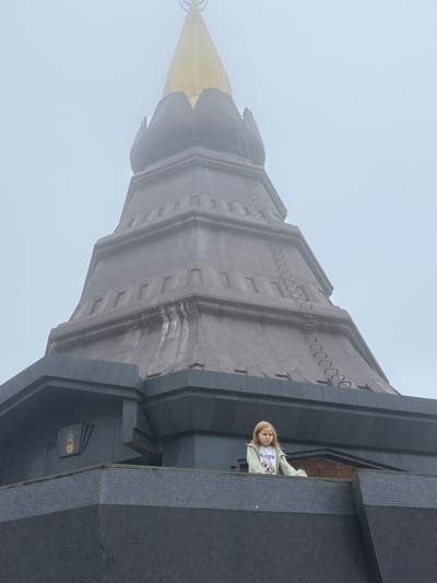 A young Caucasian girl with blonde hair stands on a tiled balcony, looking to her right. She is wearing a light-colored jacket over a white t-shirt with a graphic of two dogs. The girl is positioned in front of a large, dark, multi-tiered structure that culminates in a golden spire. The architecture appears to be a temple or monument, possibly within the city of Chiang Mai, Thailand, given its style. The background is filled with a dense fog, obscuring any further details of the surrounding environment. The lighting is overcast and diffused due to the fog.