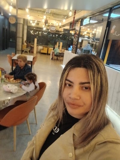 A person takes a selfie inside a well-lit establishment with high ceilings and a variety of lights. Other people sit at a table in the background. There is a chair and table in the background. The establishment looks like a restaurant, mall, or entertainment venue. The selfie taker wears a light-colored jacket and has light-colored hair.