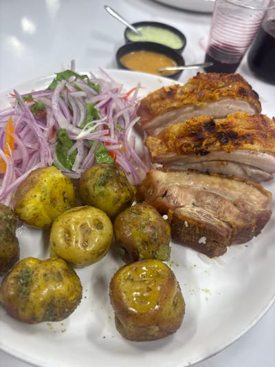 The image depicts a plate of Peruvian food, likely Chicharrón with boiled potatoes and a salad of red onion and lettuce, commonly found in La Victoria, Peru. The Chicharrón is cut into slices, revealing crispy skin and tender meat. The boiled potatoes are small and appear to have been seasoned. A salad of thinly sliced red onion and lettuce is also on the plate. Two small bowls of sauce are visible in the background, along with a partially visible glass of a dark liquid. The food is arranged on a white plate. The setting is a table, possibly in a restaurant.