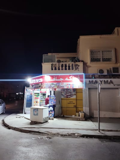 The image shows a night scene of a small convenience store, likely a "fruits secs" (dried fruits) shop, in Raoued, Tunisia. The store has a prominent red and blue neon sign that reads "FRUITS SECS" in English and Arabic script. Inside and visible through glass doors are rows of refrigerated beverages. A person wearing a red shirt is standing near the drinks section, appearing to be an employee. To the right of the store, a larger building with an awning displays the name "MAYMA" and "STOR". The street in front of the store is paved, with a concrete sidewalk and curb. Various items, including a white freezer chest, a trash can, and a broom, are visible on the sidewalk. The overall environment suggests a typical commercial street in a Tunisian town at night.