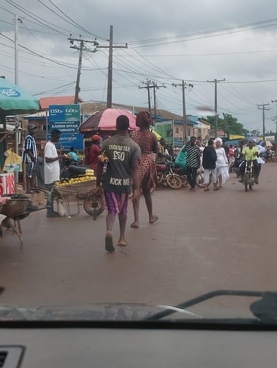 A daytime, street-level view captures a bustling scene in Aketu-Oja, Nigeria, characterized by heavy overhead electrical wires and numerous motorbikes. In the foreground, a man walks away from the camera, wearing a dark t-shirt with the text "CUCKOO TAX £50 KICK ME" and striped shorts. To his left, a woman in a leopard print dress walks in the same direction. In the middle ground, a cart filled with yellow fruits, likely oranges, is positioned near a man in a white t-shirt. Several motorbikes are parked or in motion, with people gathered around them. A person in a white wedding dress and veil is visible near the center, suggesting a wedding procession or celebration. Various stalls and shops line the background, with signage indicating businesses such as DStv and GOtv. The sky is overcast.