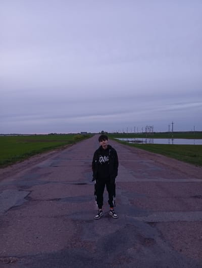 A person stands on a dilapidated asphalt road in Azyakul', Russia. The road is cracked and patched. Fields border the road on both sides, with standing water visible in the fields to the right. Utility poles line the right side of the road. The sky is overcast. The person is wearing dark clothing, including a hoodie with a graphic design, and light-colored sneakers.