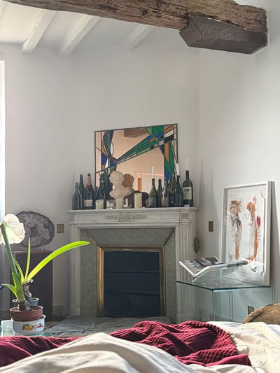 This image depicts an interior room, specifically a bedroom or living space, in Mauléon-d'Armagnac, France. The scene is illuminated by natural light entering from a window on the left.
The ceiling features exposed wooden beams, one large dark brown beam running horizontally across the top of the frame, and several smaller white painted beams perpendicular to it. The walls are uniformly white.
Centered in the image is a white marble fireplace mantle with fluted columns and a patterned gold-trimmed tile surround. The fireplace opening is dark. On the mantlepiece, a stained-glass style geometric mirror in greens, blues, oranges, and black is positioned above a white classical bust. Flanking the bust are approximately ten assorted wine and champagne bottles, including one identifiable as Moët & Chandon. Two tall white taper candles and two smaller white votive candles are also present on the mantle.
To the left of the fireplace, a dark wooden side table displays a large, dark, polished geological specimen, possibly petrified wood, on a stand. In the foreground, a potted plant resembling an amaryllis, with a prominent white flower and long green leaves, is situated in a terracotta pot resting in a decorative bowl with fruit motifs.
To the right of the fireplace, a framed vertical artwork with abstract figures in warm earth tones is leaning against the wall. Adjacent to this, a transparent glass cube table holds an open magazine or book, with a pen resting on it, possibly supported by a clear stand. The foreground reveals a bed partially covered by a white duvet and a textured dark red knitted throw blanket. No individuals are present in the scene.