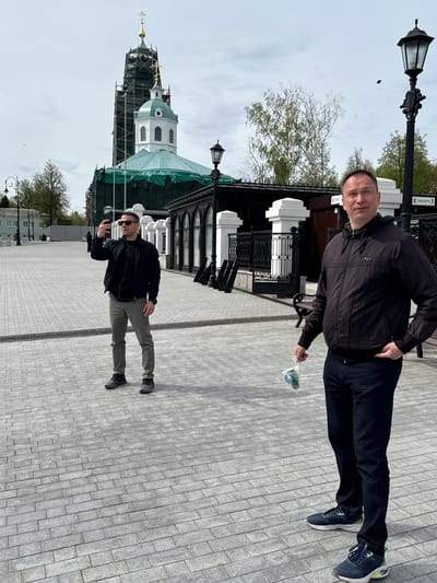 Two men stand on a paved walkway in Arzamas, Russia. A church with a green-covered dome and scaffolding is visible in the background. One man, wearing a black jacket and dark pants, holds a plastic bag with what appears to be flowers. The other man wears a dark jacket, sunglasses, and light-colored pants, holding a cell phone. Street lamps and architectural details are also present.