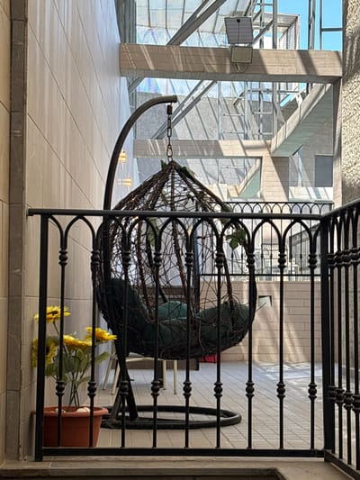 The image depicts an indoor setting with an architectural focus. The scene is characterized by the interplay of light and shadow, cast by a large, multi-story structure with exposed beams and glass elements. A large, dark metal gate or railing dominates the foreground, obscuring parts of the background.
The most prominent object in the mid-ground is a large, wire-frame basket-like structure, possibly a hanging chair or decorative element, containing a dark green cushion. It is suspended by a chain. In front of this structure, a small collection of artificial sunflowers in a terra cotta pot lies on the tiled floor. The floor itself is tiled with large, light-colored square tiles, and there are subtle decorative lights embedded in the floor in the background.
The architectural style suggests a modern building, possibly a public space like a mall, atrium, or reception area. The presence of a light fixture and a square panel mounted on a pillar in the background hints at utilities or information displays. The overall impression is one of a spacious, well-lit interior, though the time of day is not definitively ascertainable from the lighting alone. The image is oriented in landscape mode but is presented rotated 90 degrees clockwise. No people are visible. There is no visible text in the image. The context of Ayn al Basha, Jordan, implies a potentially warm climate, which could be reflected in the indoor design choices.