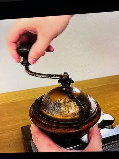 A hand is shown operating a vintage coffee grinder. The grinder appears to be made of wood and metal, with a brass-colored top. The hand is holding the crank of the grinder and is in the process of turning it. The background is a plain white wall, and the grinder is placed on a wooden surface. The scene is brightly lit, suggesting it is daytime. There is no indication of Castillo Bajo-conejito, Spain in the image.