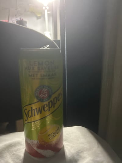 A can of Schweppes Lemon is standing upright on a white surface. The can has a yellow and green design with the text "LEMON AUX SAVEURS MET SMAAK" and "Schweppes". There is also a depiction of a tennis ball with "ROLAND GARROS" written on it. The background is dark and out of focus, with a bright light source on the left. The scene is indoors, and no specific location in Carrières-sous-Poissy, France, can be determined.