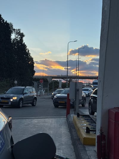 The image depicts an outdoor urban scene at sunset, specifically at a gas station adjacent to a road junction in Pardes Hana - Karkur, Israel.

In the immediate foreground, the fuel cap of a dark vehicle is open, with a fuel nozzle inserted, indicating refueling. A partial yellow Israeli license plate is visible on this vehicle. To the right, a red fire extinguisher is mounted on a light-colored structural pillar of the gas station, with yellow and black ground barriers visible below.

The midground features a multi-lane asphalt road with active vehicular traffic. A silver-grey Skoda Yeti SUV, bearing Israeli license plate 65-077-73, is positioned in the left lane with its headlights illuminated, appearing to turn left. A dark SUV, with Israeli license plate 918-10-003, is present in the right lane, facing away from the viewer. Additional vehicles are visible further down the road, indicating traffic congestion. A red circular 'no entry' sign is visible near industrial piping to the left of the road. Multiple dark-grey streetlights line the road.

In the background, a large concrete and metal bridge/overpass spans horizontally across the scene. To the far left, a dense line of tall, dark green trees borders the road. The sky displays a sunset with distinct crepuscular rays emanating through clouds, casting orange and yellow light against a light blue upper sky. No individuals are discernible in the frame.