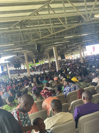 The image shows a large crowd of people seated in rows of chairs inside a large, open-air structure with a complex metal framework supporting the roof. The people are diverse in age and clothing, many wearing colorful traditional attire. The scene appears to be an event or gathering taking place in a sizable venue in Otta, Nigeria. Visible above the crowd are speakers hanging from the ceiling. The image is shot from a high vantage point along one of the sides of the venue.