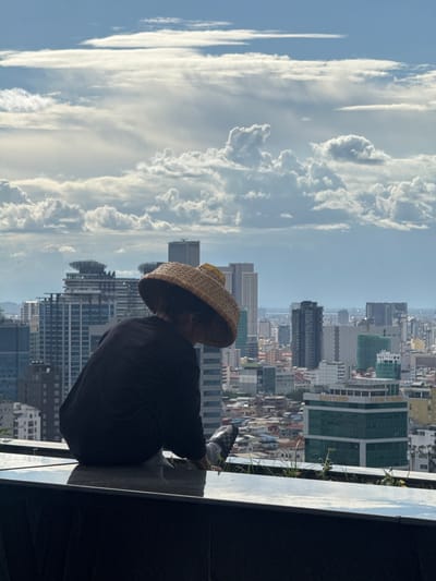 A person wearing a conical hat and black clothing sits on a reflective surface overlooking a sprawling cityscape under a cloudy sky. The person is facing away from the camera, looking towards the buildings. The city appears dense with a mix of modern high-rise buildings and smaller structures. The sky is filled with dramatic clouds. The foreground shows a dark, possibly stone, ledge with some sparse greenery growing from it. The overall impression is one of quiet contemplation or observation amidst an urban environment. The location is identified as Khan Boeng Keng Kang, Cambodia.
