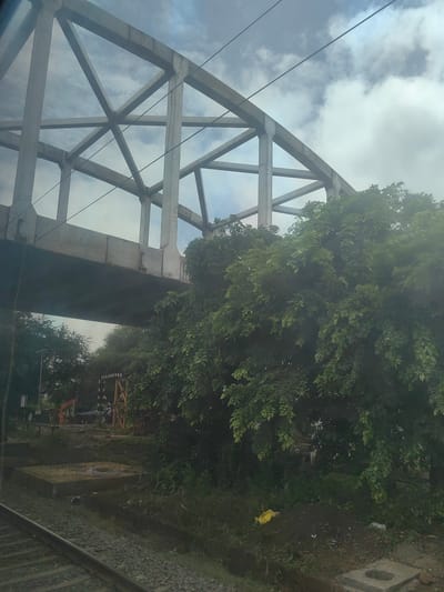 The image depicts a railway track running diagonally from the lower left corner towards the center. Overhead, a large concrete bridge with a complex truss structure spans across the frame. Dense green foliage of trees obscures the lower portion of the bridge and the surrounding area, suggesting a lush environment. In the background, under the bridge, several construction elements are visible, including what appears to be an orange construction vehicle and a wooden structure with black and white markings, possibly a signal or marker. The ground is uneven and appears to be a mix of dirt, gravel, and some construction debris. A yellow plastic bag is visible on the ground amongst the vegetation. The sky above is cloudy with patches of blue visible through the clouds. The overall scene suggests an outdoor location, possibly on the outskirts of Kamtara, India, where infrastructure development or maintenance might be underway near a railway line.