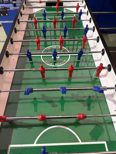 The image shows a close-up, angled view of a foosball table. The table features red and blue players affixed to chrome rods that extend across the playing surface. The rods are spaced evenly, and some have springs at the top. The playing field is green, with white lines marking the boundaries and center circle of a soccer field, along with "SOCCER - BALL" written on the center circle. The table's side is visible, showing a blue and white color scheme. The location is not discernible from the image itself. There are no people present.