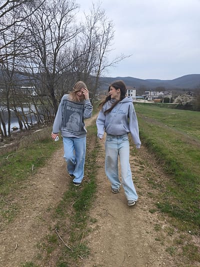 Two young women walk side-by-side on a dirt path lined with grass in Орлиновский округ, Ukraine. The woman on the left has blonde hair, and wears a gray sweatshirt with the word "Blondie" on it, light blue jeans, and black shoes. She holds a phone in her left hand and touches her head with her right. The woman on the right has brown hair and wears a gray hooded sweatshirt with the words "Never Look Back" on it, light blue jeans, and gray shoes. She looks towards the woman on the left. To the left of the path are bare trees and a body of water. To the right is a grassy field and houses in the distance. Mountains are visible in the background under a cloudy sky.