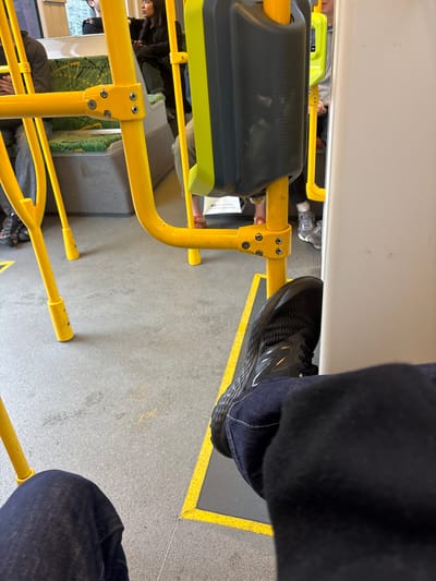 An interior view from a tram in Brunswick East, Australia, shows passengers seated on green and white patterned upholstery. Yellow metal poles and handrails dominate the frame, with a gray panel and a yellow-green indicator light visible near the center. The floor is a concrete-like gray surface marked with yellow safety lines. In the foreground, a person's legs are visible, wearing dark blue jeans and black athletic shoes. Another passenger's legs, wearing gray sneakers, are seen further back. The lighting is artificial, typical of a public transport vehicle.