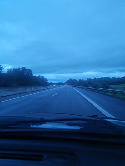 The image is a point-of-view shot from inside a vehicle traveling on a highway at dusk or during overcast conditions, likely near Liptovský Mikuláš, Slovakia, based on the provided context. The highway is wet, indicating recent rain. The road has multiple lanes marked by white lines. On the left side of the highway, there is a concrete barrier and then a dense line of trees. On the right side, there is a metal guardrail and then a grassy embankment leading to more trees and hills in the distance. The sky is heavily overcast with dark, textured clouds. In the distance, on the highway, a truck with illuminated red taillights is visible. The lower part of the image shows the dashboard and windshield of the vehicle. The overall lighting is dim and blue-toned.