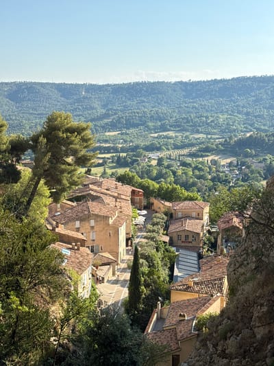 This is a high-angle, medium shot of a village nestled in a valley, with rolling hills and lush green trees in the background. The village features buildings with terracotta tile roofs and light-colored walls, characteristic of Moustiers-Sainte-Marie, France. In the foreground, a large pine tree leans into the frame from the left, with its branches obscuring some of the village's rooftops. The buildings are densely packed, with narrow streets winding between them. Some buildings have awnings or outdoor seating areas, suggesting commercial establishments. A few people are visible walking on the streets. The sky is clear and blue, indicating a sunny day. The overall impression is of a charming, sun-drenched village in a picturesque natural setting.