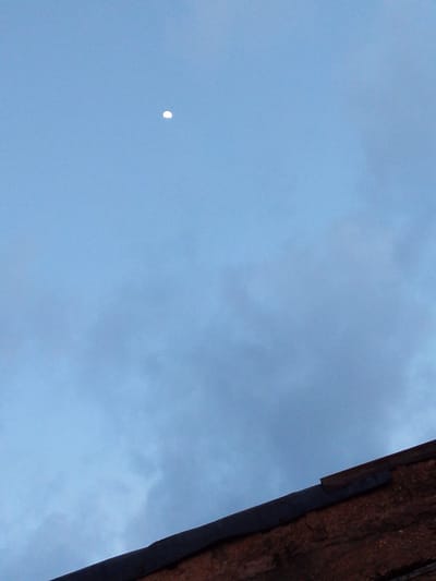 The image captures a scene looking upwards towards a pale blue sky, with some soft, wispy clouds visible. A sliver of the moon is present in the upper portion of the frame, appearing as a bright crescent against the twilight sky. In the foreground, the edge of a dark, sloped roof made of reddish-brown, textured material is visible, suggesting an outdoor setting, possibly residential. The lighting indicates it is either early morning or late evening, given the soft illumination and the visible moon. There is no text or discernible activity taking place, creating a serene and quiet atmosphere. The overall impression is one of a calm, atmospheric moment in what appears to be a Nigerian setting.