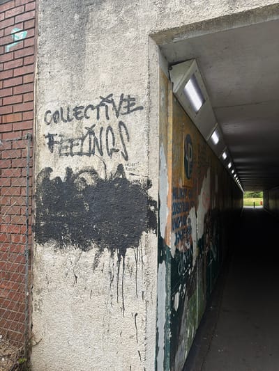 The image shows the interior of a tunnel or underpass in Queensferry, United Kingdom. The walls are concrete, and the floor is a series of panels that appear to have old posters or advertisements affixed to them. A row of rectangular lights is mounted along the left wall. Graffiti, including the words "collective feeling", is visible on the lower portion of the right wall, partially covered by a large black blotch. A brick walkway and a wire mesh fence are at the bottom of the frame. The tunnel extends into the distance, with an opening showing green foliage at the far end.