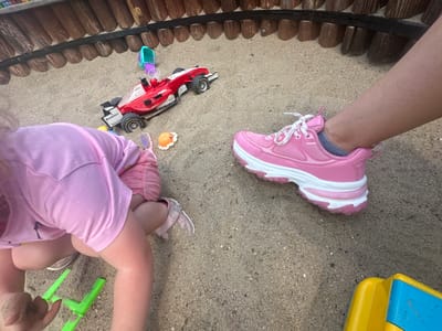 The image depicts a sandbox in the city of Donchovci, Bulgaria, where a young child is playing. The child, wearing a pink shirt and shorts, is kneeling in the sand, engaged with a green toy shovel. Nearby, a red toy race car with the word "BIONERGY" on its side is positioned on the sand. Several other small toys are scattered around, including a purple brush, an orange seashell-shaped toy, and a teal and purple toy. To the right of the child, an adult's leg is visible, clad in a bright pink and white chunky sneaker. The background features a wooden fence.