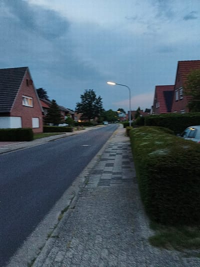The image depicts a quiet street in Papenburg, Germany, at dusk or twilight, with overcast skies. The street is paved, with a concrete sidewalk running along the right side. The sidewalk has a pattern of rectangular and square paving stones, some of which are darker than others. A low, neatly trimmed green hedge borders the sidewalk on the right.
On the left side of the street, there are residential houses. The closest house is made of red brick with a dark tiled roof. It has at least two windows visible, one of which is lit from within. The house is also bordered by a green hedge. Further down the street, more houses and trees are visible, along with a streetlamp that is illuminated. A white car is parked in front of one of the houses.
The overall impression is of a peaceful suburban environment. There are no people or animals visible in the image, and no actions or interactions are taking place. The scene is static and serene. The image has a slightly grainy texture, consistent with low-light photography.