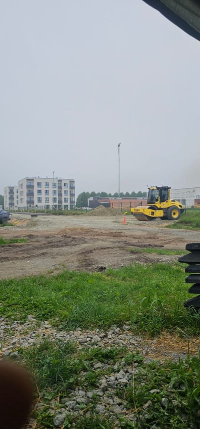 The image depicts a construction site in Maardu, Estonia. In the background, a modern multi-story apartment building with a light-colored facade and balconies is visible. To the right of the apartment building, a brick structure is present. In the middle ground, a bright yellow Bomag vibratory roller is parked on a gravel surface, with piles of gravel and a bright orange traffic cone nearby. The ground in front of the roller is uneven and shows tire tracks from construction vehicles, indicating ongoing work. The foreground is filled with patches of green grass and loose gravel. The sky is overcast, suggesting a cloudy day.