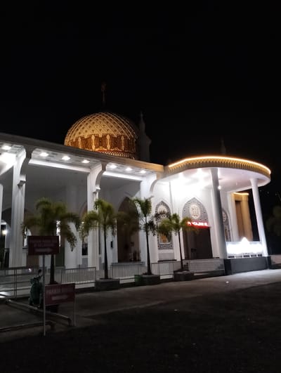 The image shows the exterior of a mosque at night in Blangmetimu, Indonesia. The mosque features a prominent golden dome with intricate patterns, illuminated by spotlights. The facade is white with arched walkways supported by columns. Palm trees are planted in front of the building. Red signs indicating parking areas are visible, one for motorcycles ("PARKIR RODA DUA DI BELAKANG") with an arrow pointing to the right, and another for cars ("PARKIR MOBIL") also with a right-pointing arrow. A green scooter is parked near the motorcycle parking sign. The ground in front of the mosque is paved with dark asphalt. The sky is dark, indicating it is nighttime, and subtle lighting outlines the upper edges of the mosque's structure.