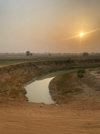 An expansive rural landscape bathed in golden sunlight. A small body of water reflects the hazy sky. A barren stretch of land contrasts with a strip of vegetation. A road runs alongside the water. The sun dominates the sky, casting long shadows and illuminating the earth.