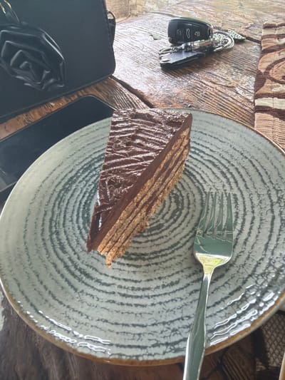 The image presents a high-angle, close-up view of a dessert setting on a wooden table, likely in Sushina, Bulgaria. The central focus is a slice of layered chocolate cake placed on a light grey ceramic plate with a unique concentric circular pattern in darker grey. The cake displays multiple thin layers of sponge or wafer interspersed with thick, rich chocolate cream or ganache, topped with a textured chocolate glaze. A silver four-tine fork rests on the plate to the right of the cake.

To the left of the plate, a set of car keys is visible on the wooden surface. The keyring holds a black car remote fob, displaying standard lock, unlock, and trunk release buttons, along with a metallic traditional key. Below the keys, a part of a black wallet or small bag is seen, adorned with a delicate black rose-shaped embellishment.

The table itself is made of wood, exhibiting a pronounced grain and slightly worn texture. Portions of a brown and beige patterned fabric, possibly a placemat or part of a larger textile, are visible in the top left and top right corners of the frame. The lighting indicates daytime, with reflections visible on the metallic fork and the cake's glaze, suggesting a bright environment. The scene captures a moment of leisure, perhaps a snack or dessert break. No discernible text is visible other than the symbols on the car key fob.